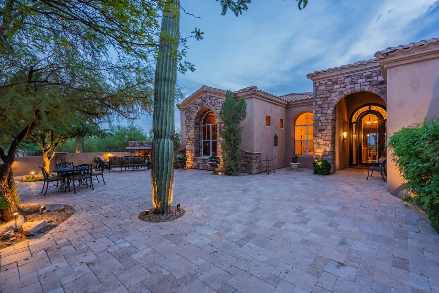 Arizona home at twilight with a stone arched entry, glowing windows, a lit saguaro and a paved courtyard with outdoor dining and a fireplace